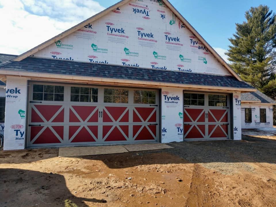 Two-car garage with red cross-style doors, gray trim, and windows, under construction, covered in Tyvek.