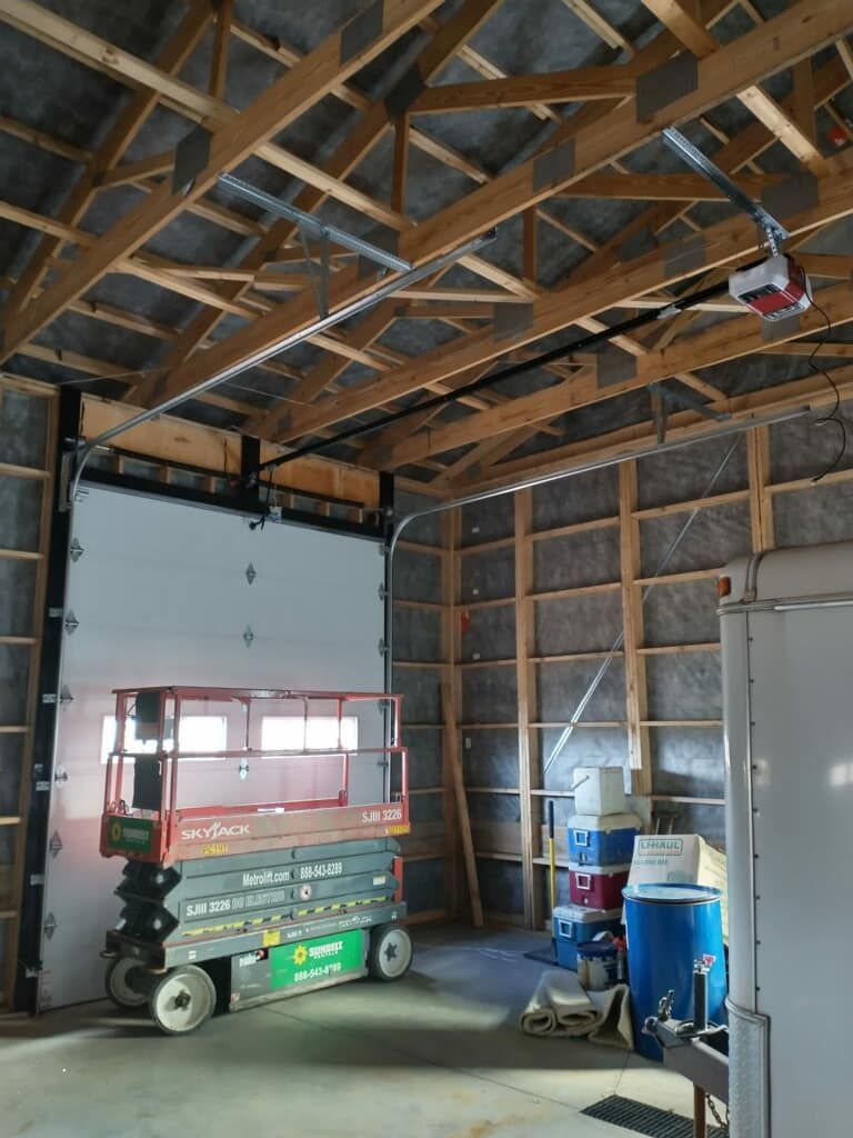 Inside a workshop, a large closed white garage door with a scissor lift in front; wood beams and a garage door opener above.