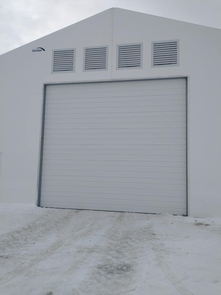 White building with closed garage door, snow-covered ground in front. Four vents above the door.