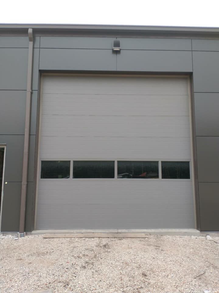 Gray industrial building with a large closed garage door, four windows, and gravel ground.