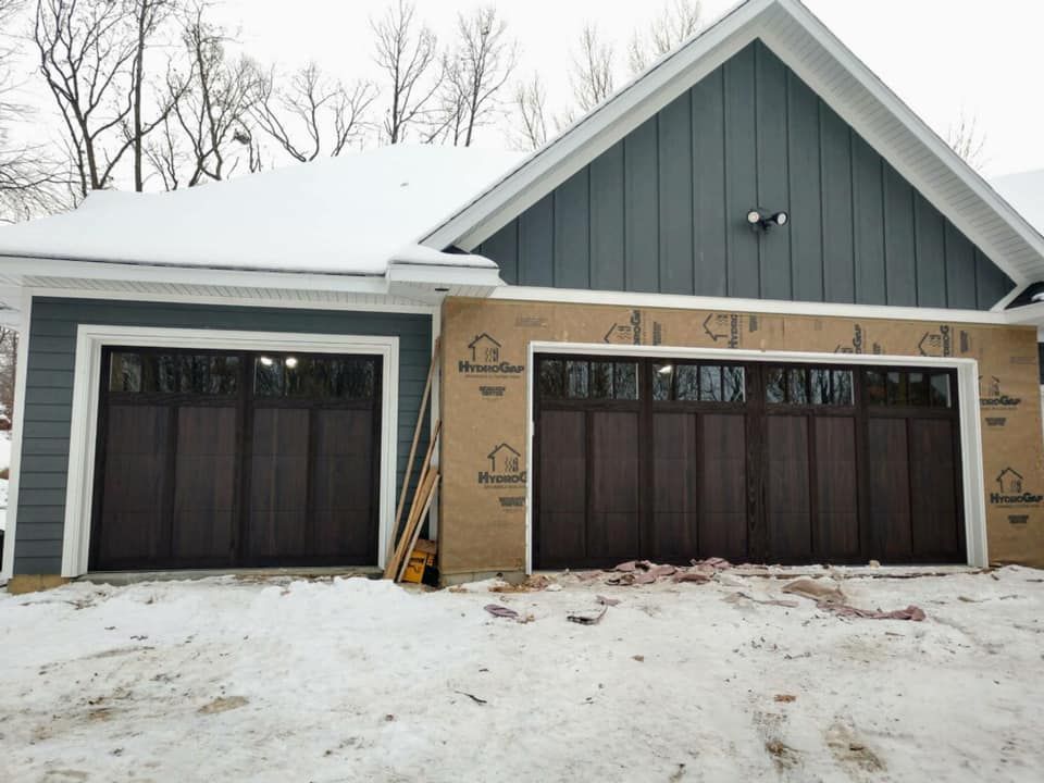 Two-car garage with brown doors and gray siding, one side under construction. Snow covers the ground and roof.