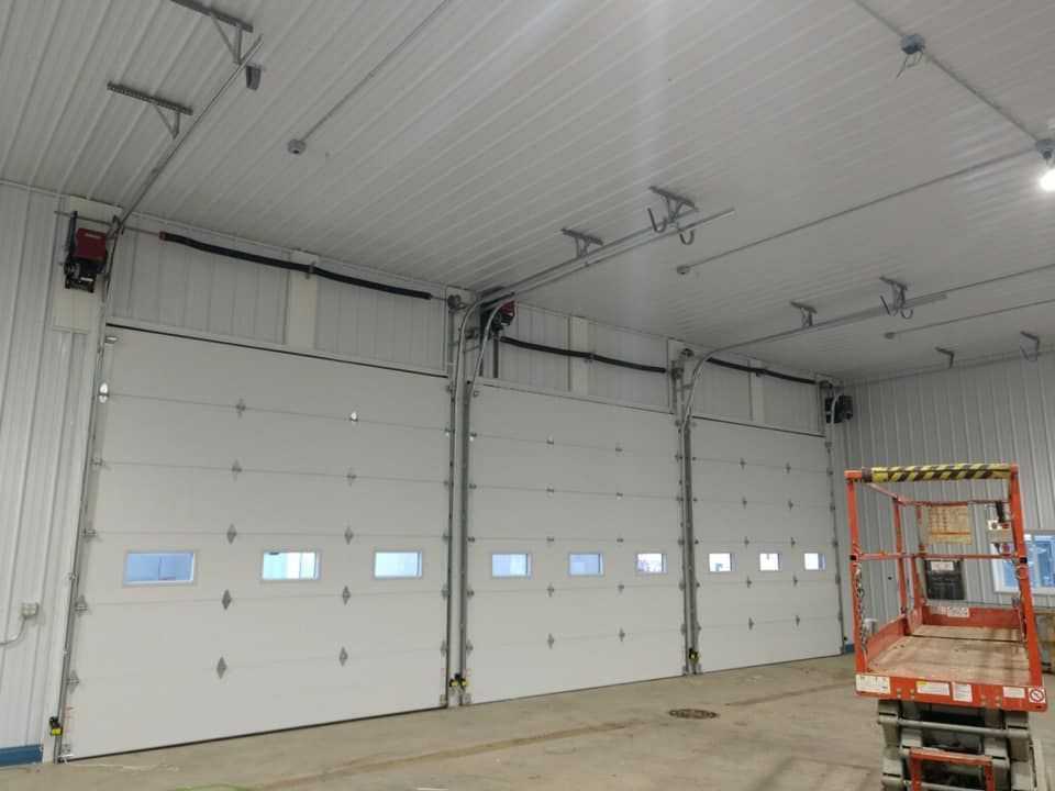 Three white overhead garage doors inside a large building. A scissor lift is on the right.