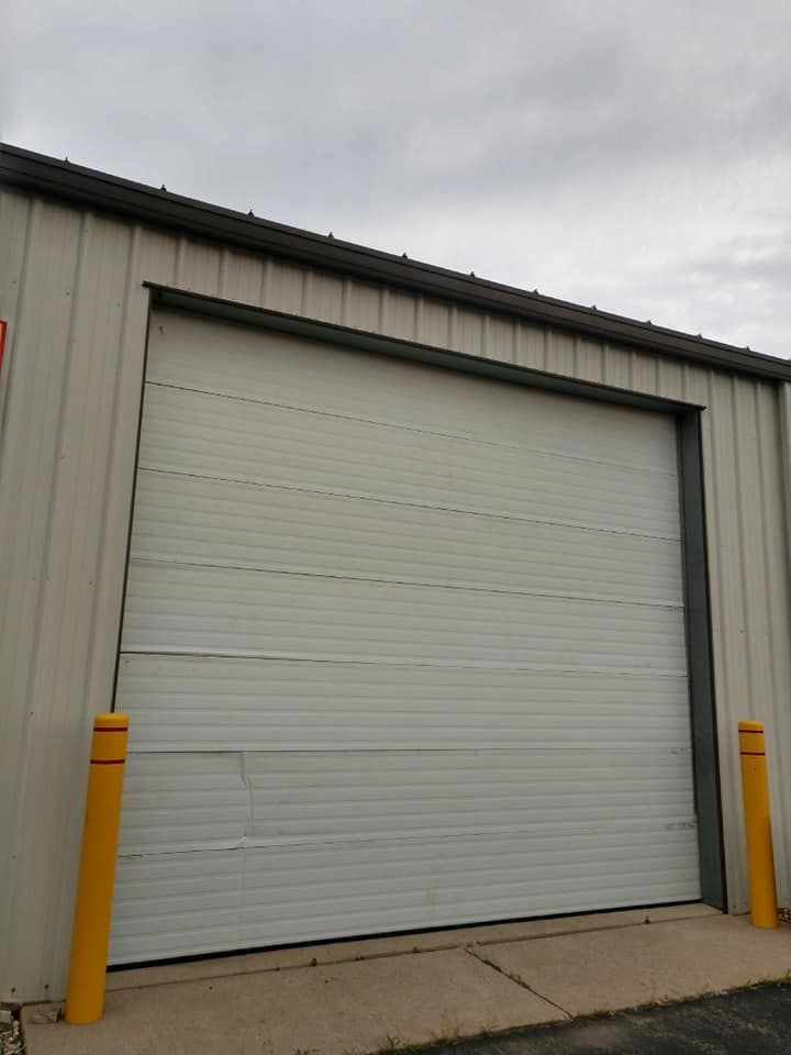 White overhead garage door with minor dents in a metal building, yellow bollards on either side.