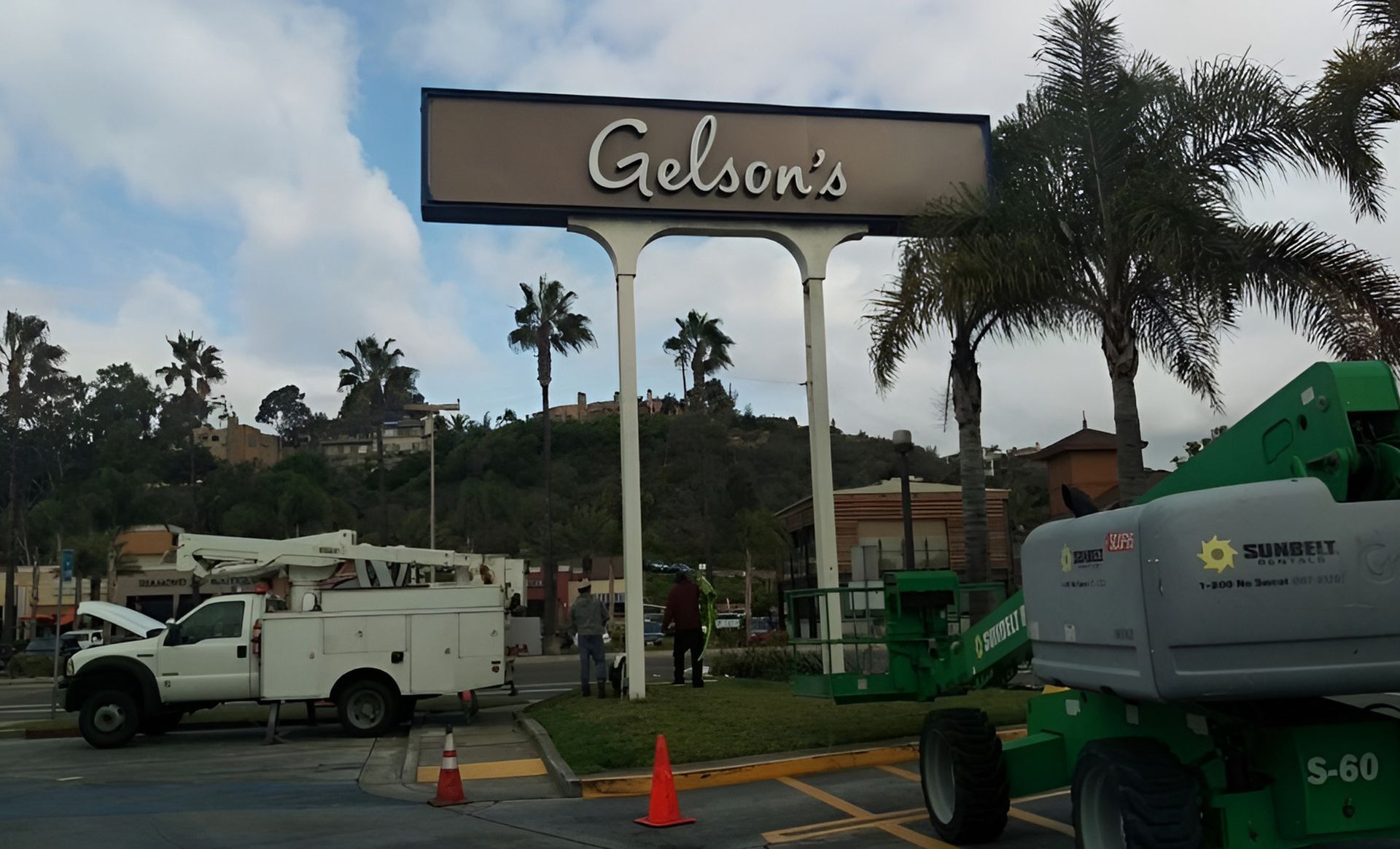 Gelson's sign at a roadside entrance with palm trees, utility trucks, and orange cones