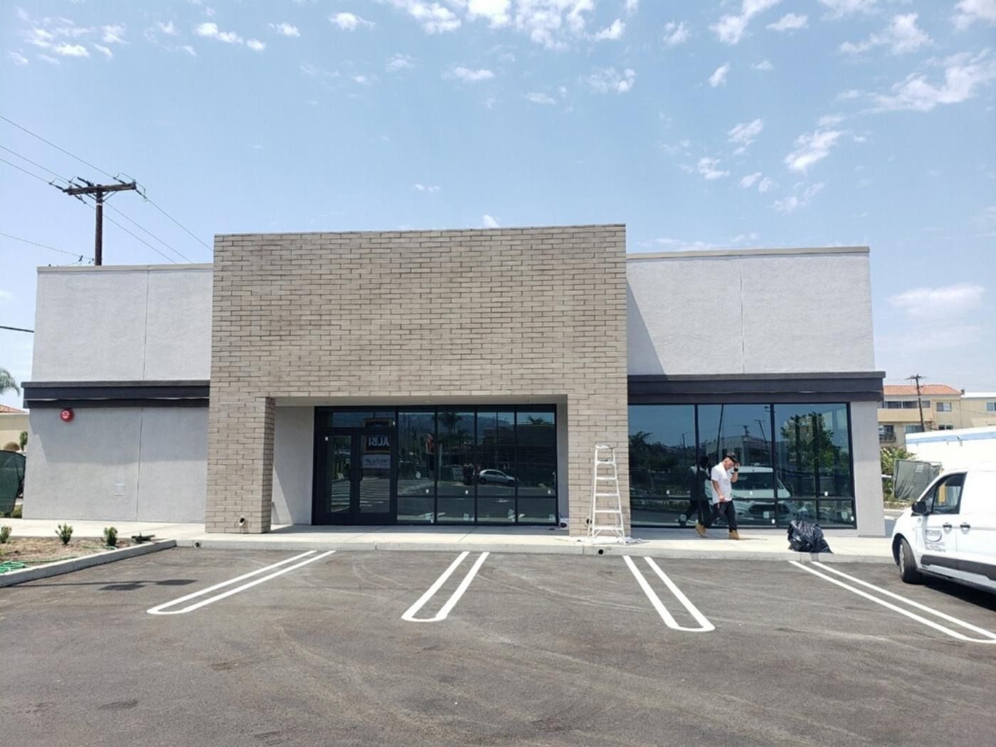 Exterior of a commercial building with glass front, under construction. Man working on the right side, parking lot in front.