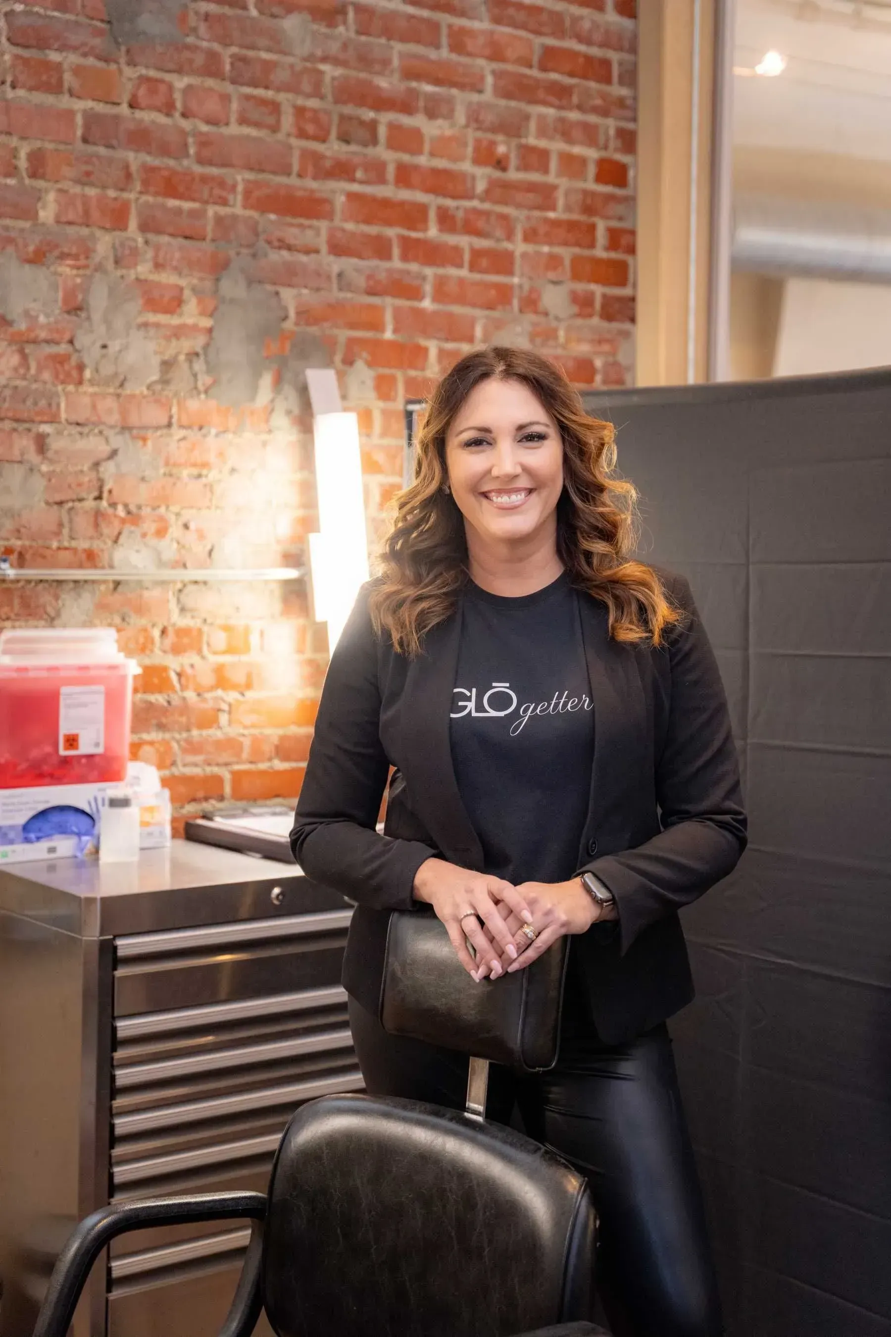 Woman standing by a salon chair in a brick-walled room, smiling with folded hands.