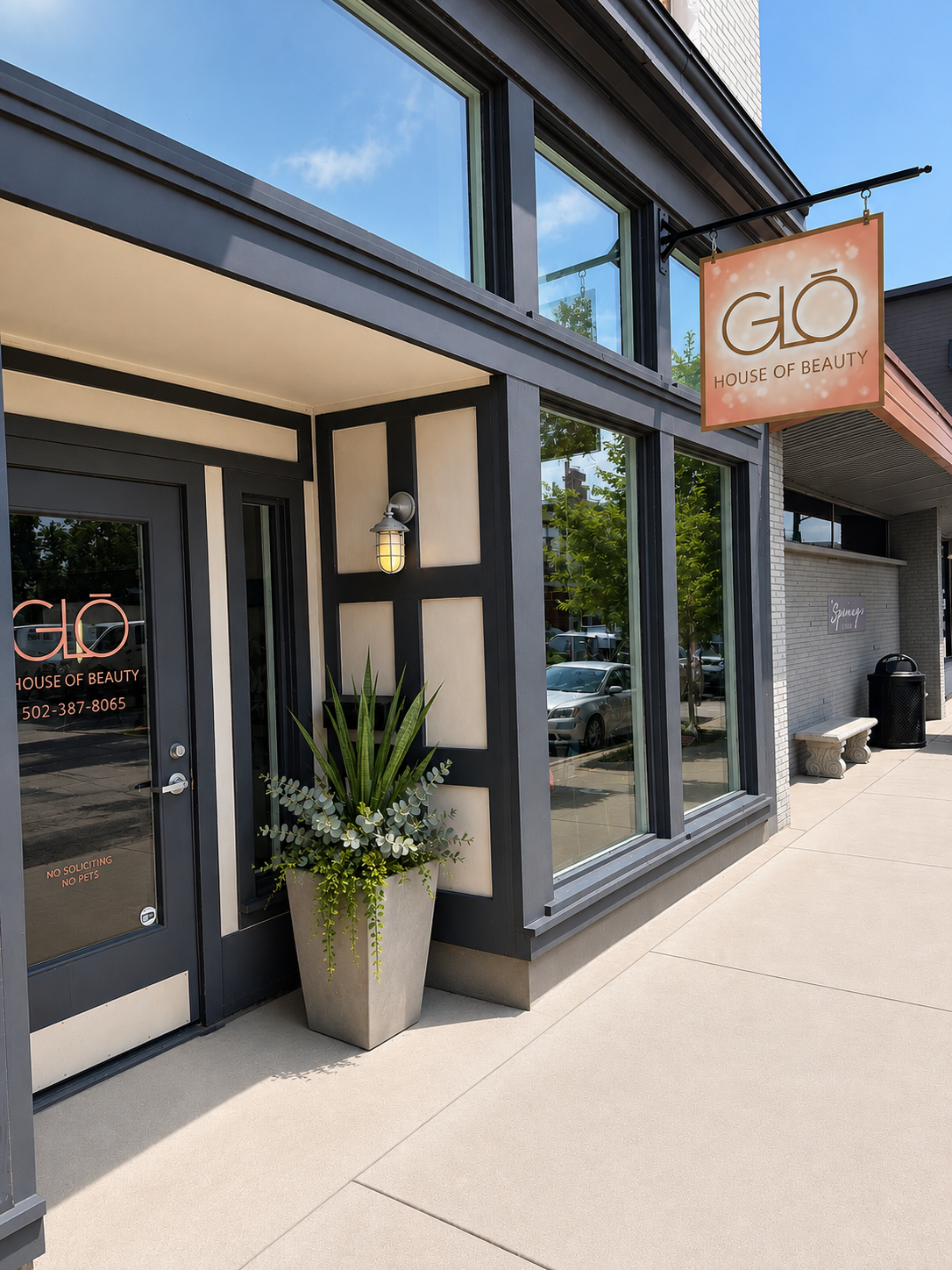 Storefront with large windows, black trim, potted plant, and hanging orange “Glo” sign on a sunny sidewalk.
