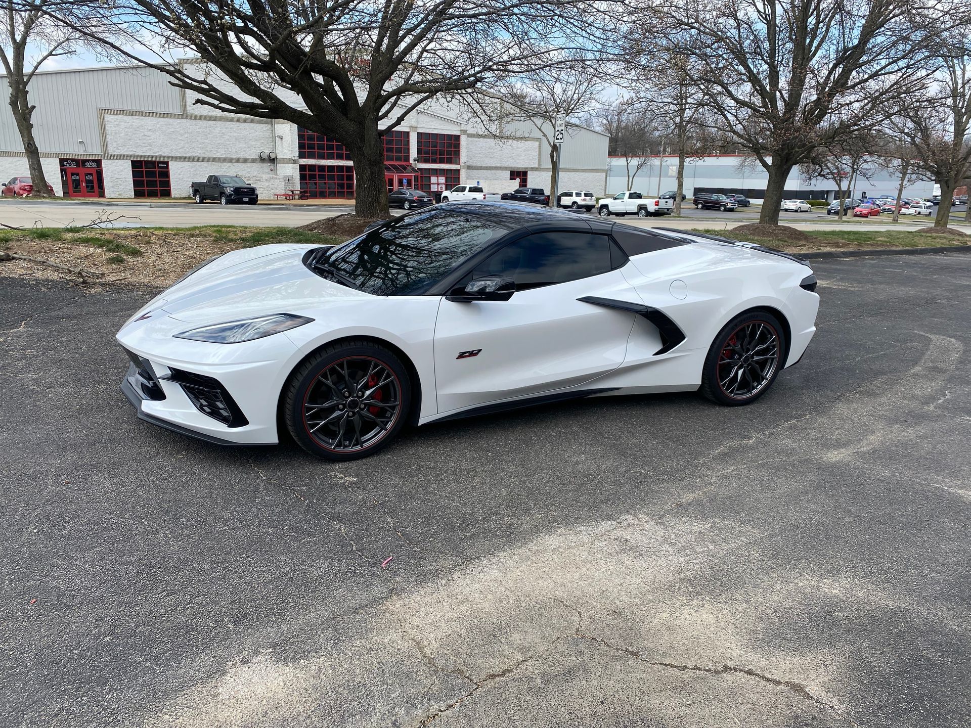 White sports car parked on asphalt, black wheels, dark roof, in front of buildings.