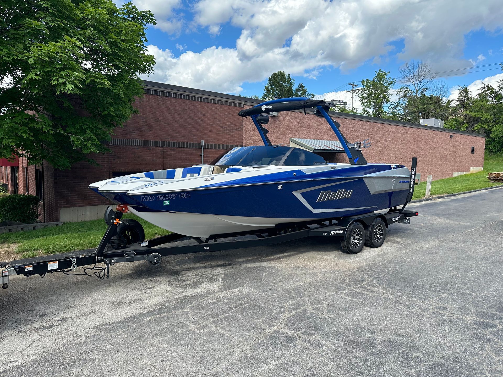 Blue and white wakeboard boat on a trailer parked outside a brick building.