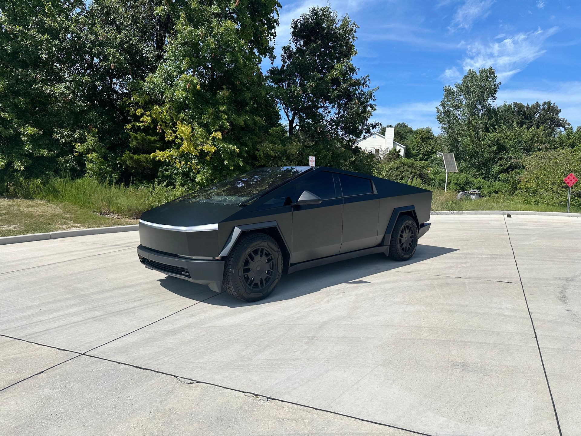 Tesla Cybertruck parked on a concrete surface, black body, dark wheels, trees and blue sky in background.