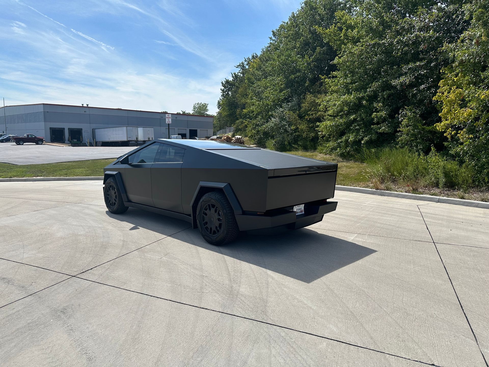 Tesla Cybertruck, matte black, parked on pavement, sunny day. Trees and buildings in background.