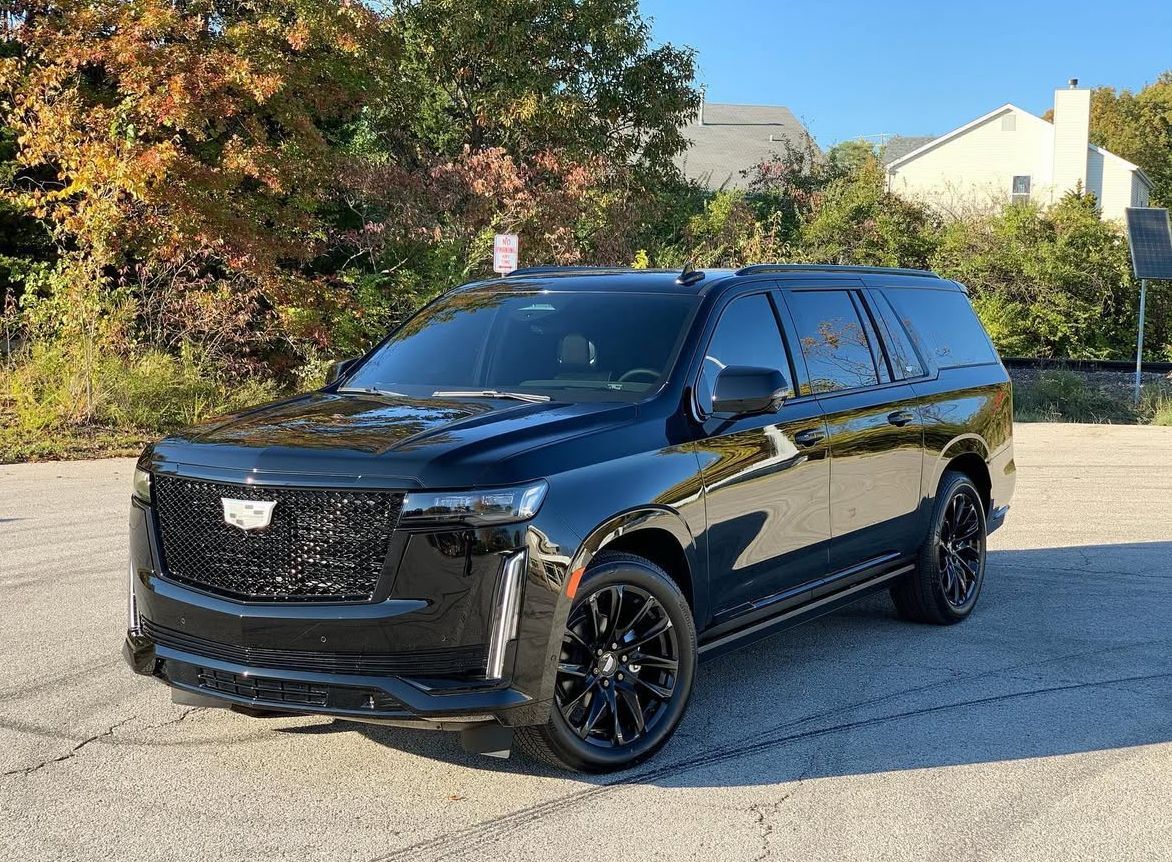Black Cadillac Escalade SUV parked on asphalt, trees and building in the background.