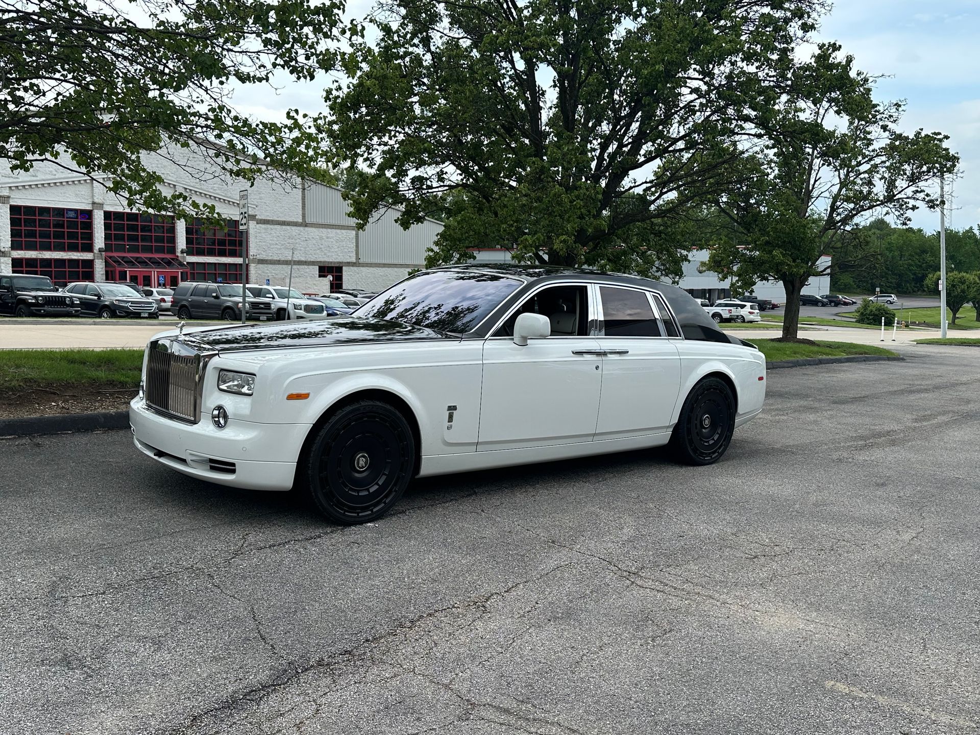 White Rolls-Royce Phantom with black accents parked on gravel. Green trees and a building in the background.