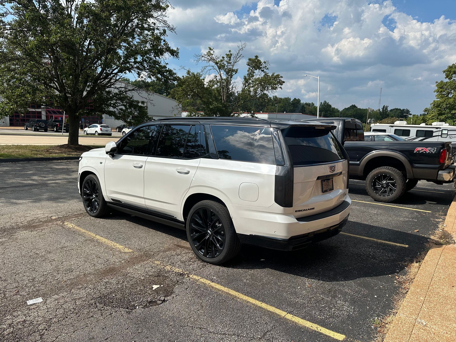 White SUV parked in a lot, black rims, tinted windows. Dark colored truck parked next to it.