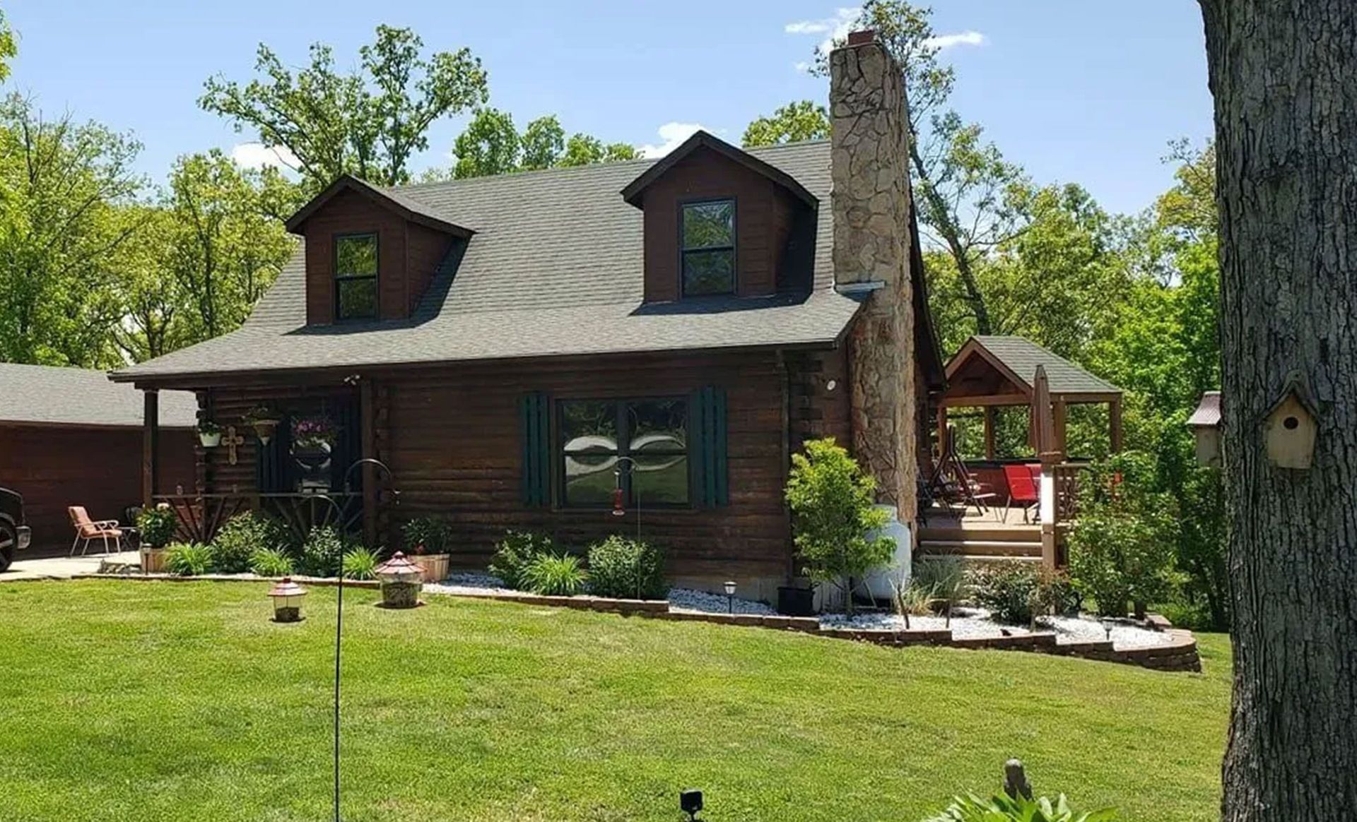 Log cabin with a stone chimney, green lawn, and trees.