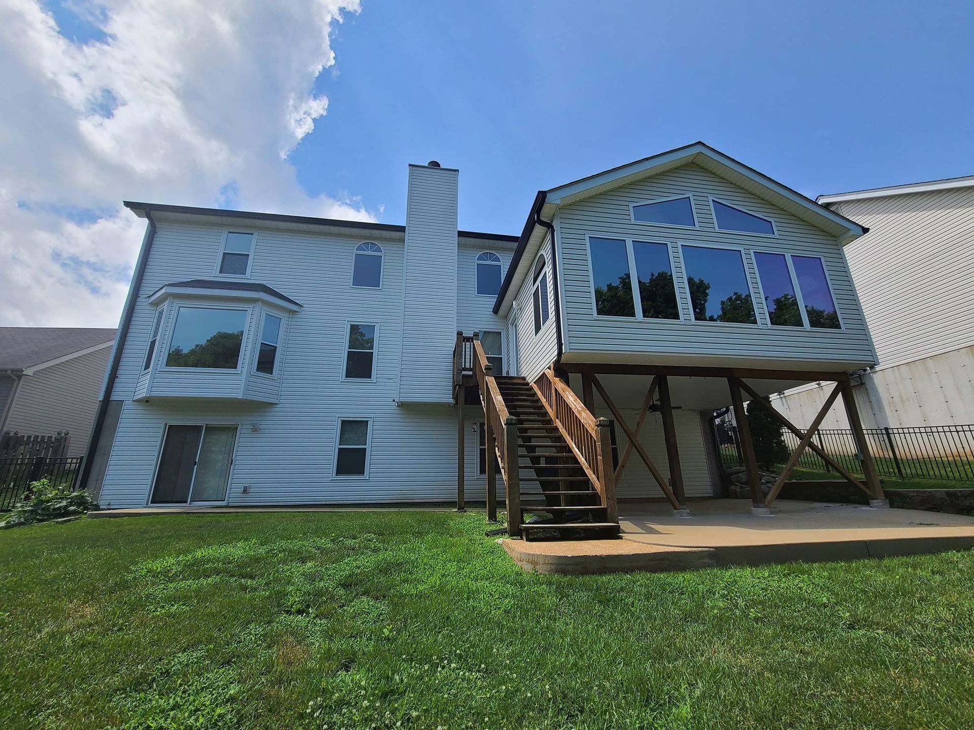 Back view of a light-colored two-story house with a bay window, sunroom, chimney, and wooden stairs, on a grassy lawn under a blue sky.