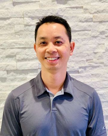 A man in a gray shirt is smiling in front of a stone wall.