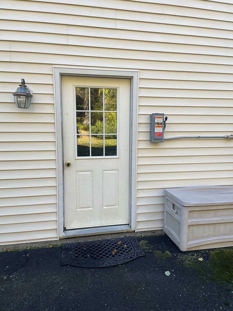 Weathered, light gray door with glass panes, framed in light gray, set in a light yellow siding wall; a black mat, and a white storage box. - after