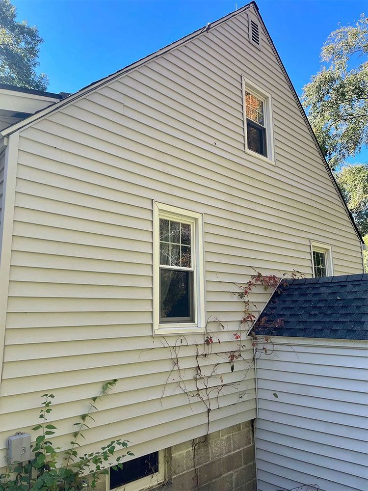Side view of a weathered light-colored house with multiple windows and visible wood siding. A smaller building is attached. - after