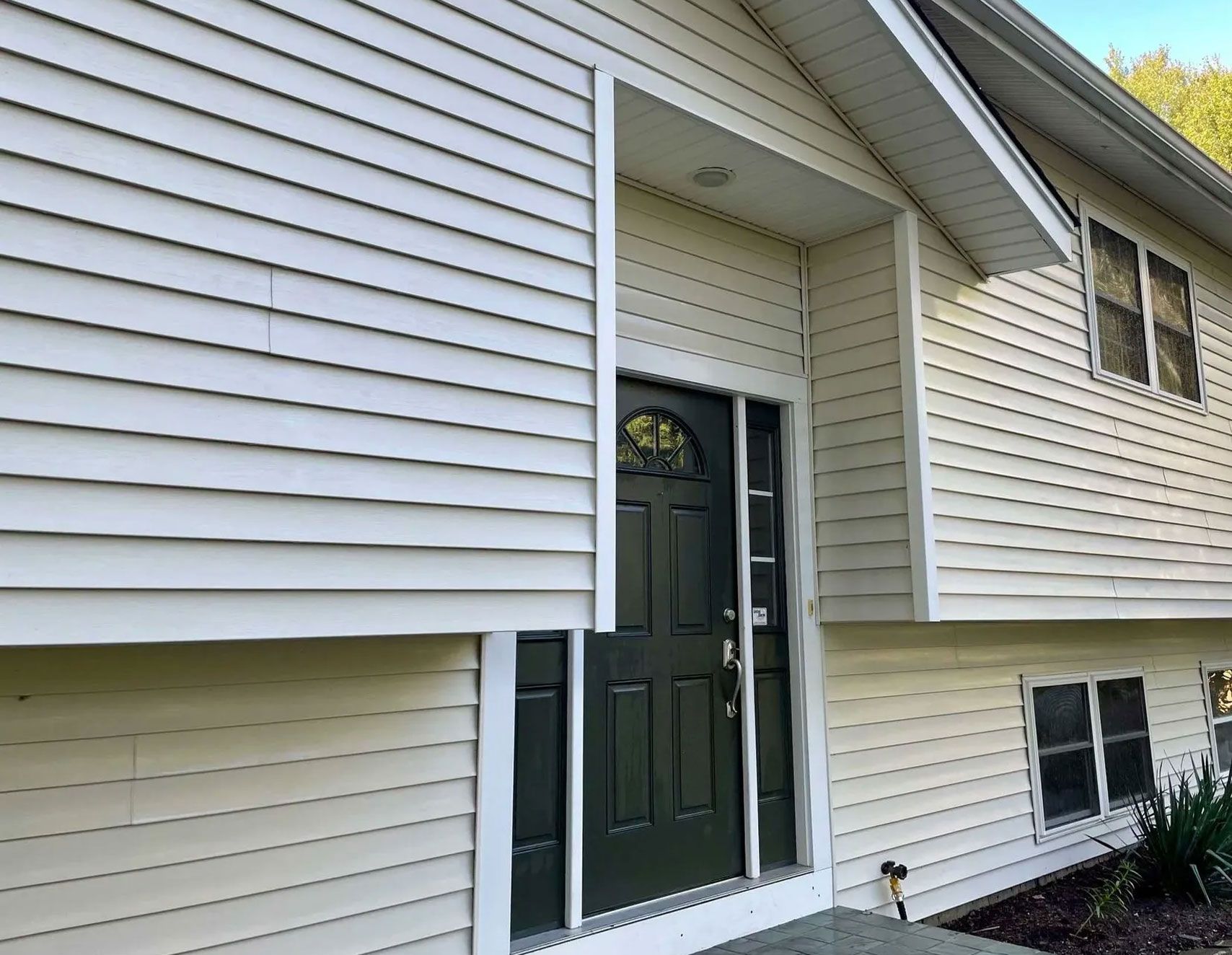 Off-white house with siding and dark door, covered in green mildew. Blue sky - after