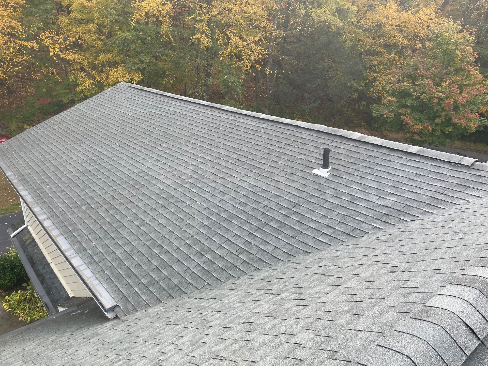 Dark shingled roof with a vent, viewed from a higher roof. Fall foliage in the background. - after