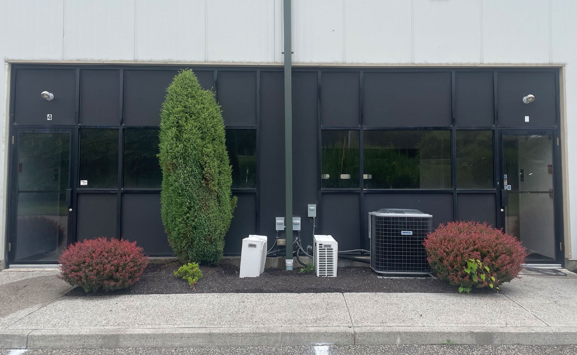 Exterior of a building with dark-framed windows, shrubbery, and a central evergreen tree. An AC unit sits on the right. - after