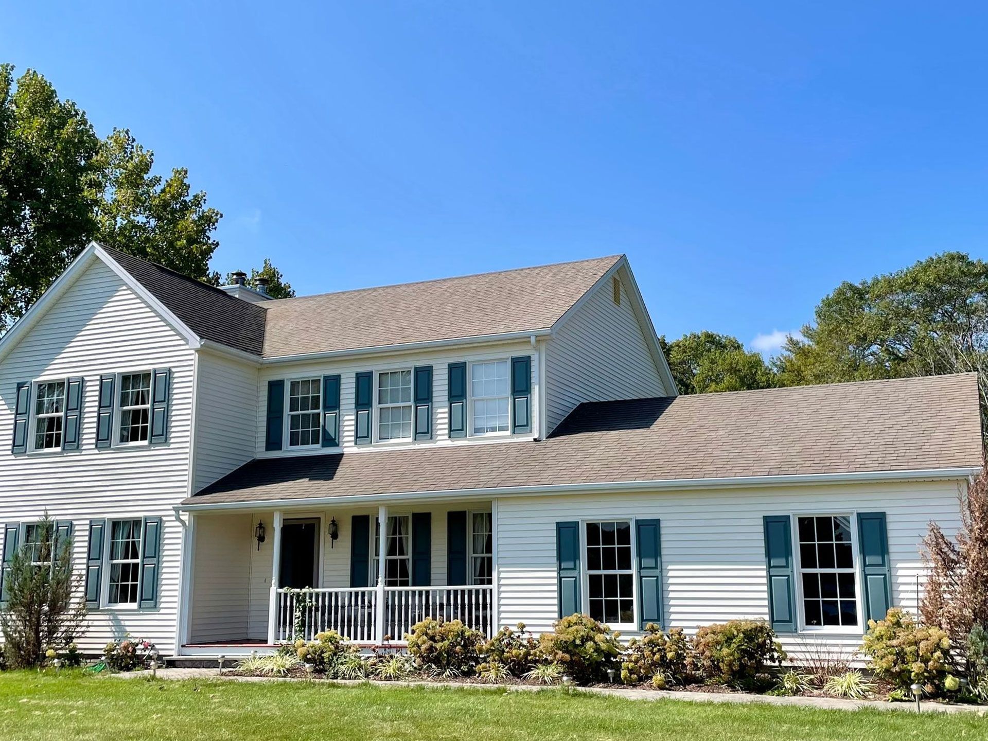 Two-story beige house with green shutters and brown roof under a blue sky. - after