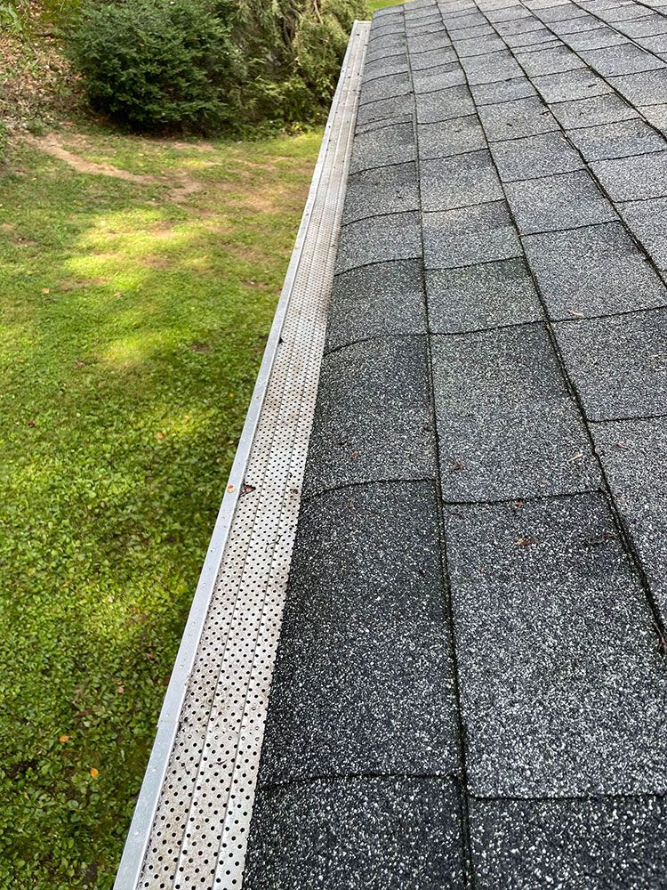 Gutter clogged with leaves and debris alongside a roof with gray shingles. Green grass in the background - after