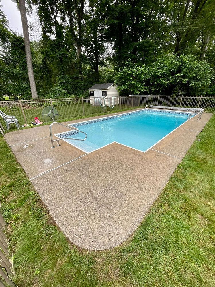 An outdoor swimming pool surrounded by concrete, grass, and a wooden fence. The pool water is blue, - after