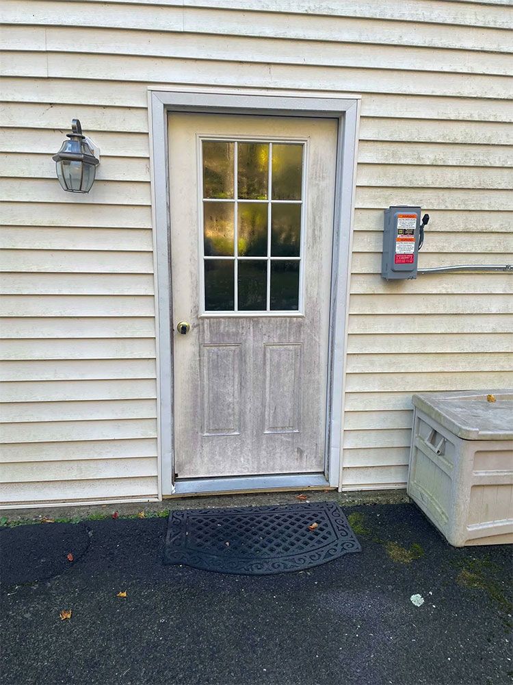 Weathered, light gray door with glass panes, framed in light gray, set in a light yellow siding wall; a black mat, and a white storage box. - before