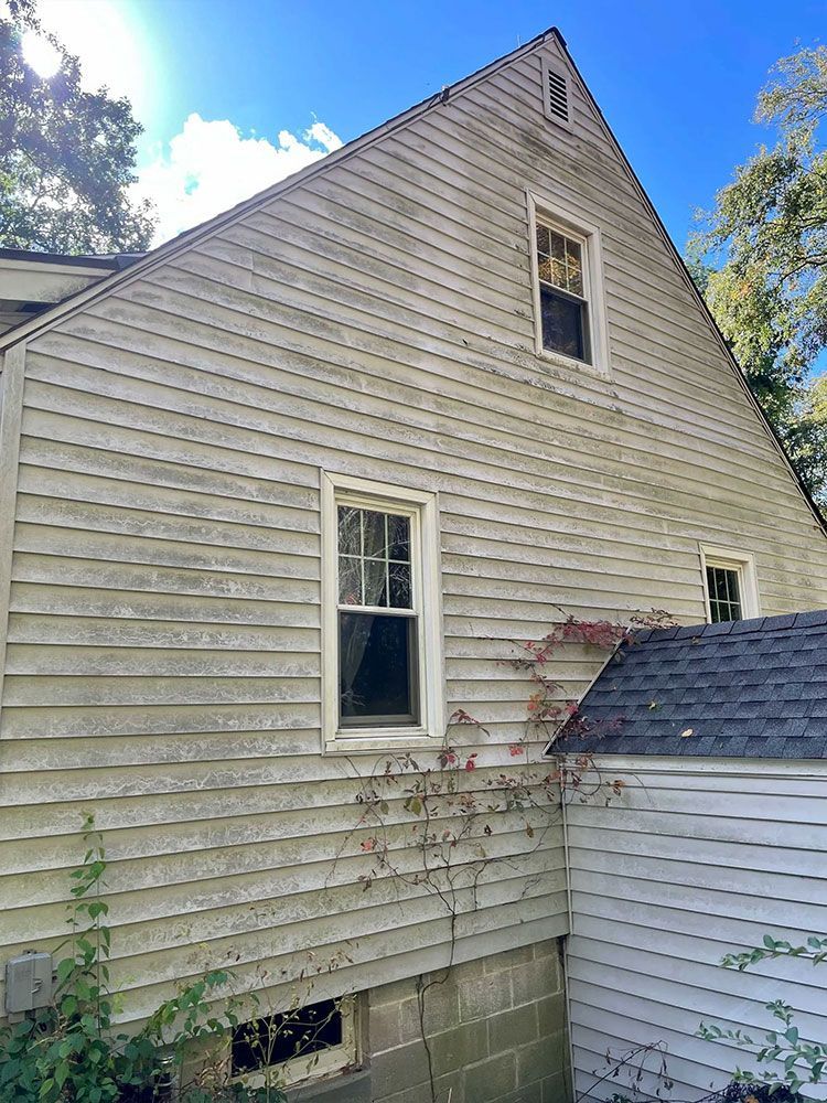 Side view of a weathered light-colored house with multiple windows and visible wood siding. A smaller building is attached. - before
