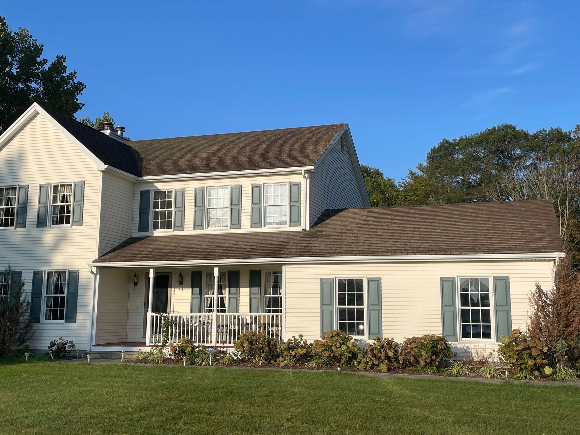 Two-story beige house with green shutters and brown roof under a blue sky. - before
