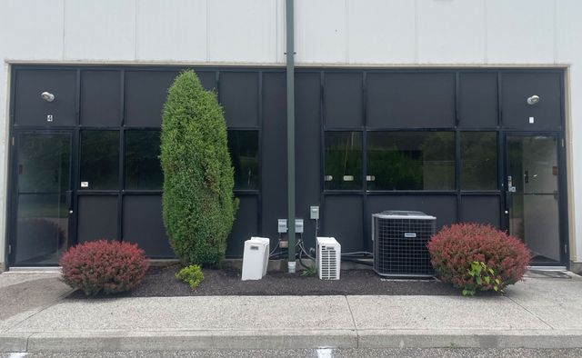 Exterior of a building with dark windows and doors, landscaping includes bushes and a tree.