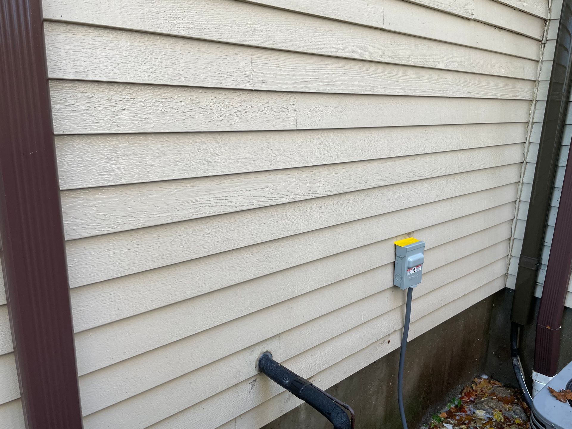 Beige siding on a house exterior with a brown gutter, electrical box, and black pipe.