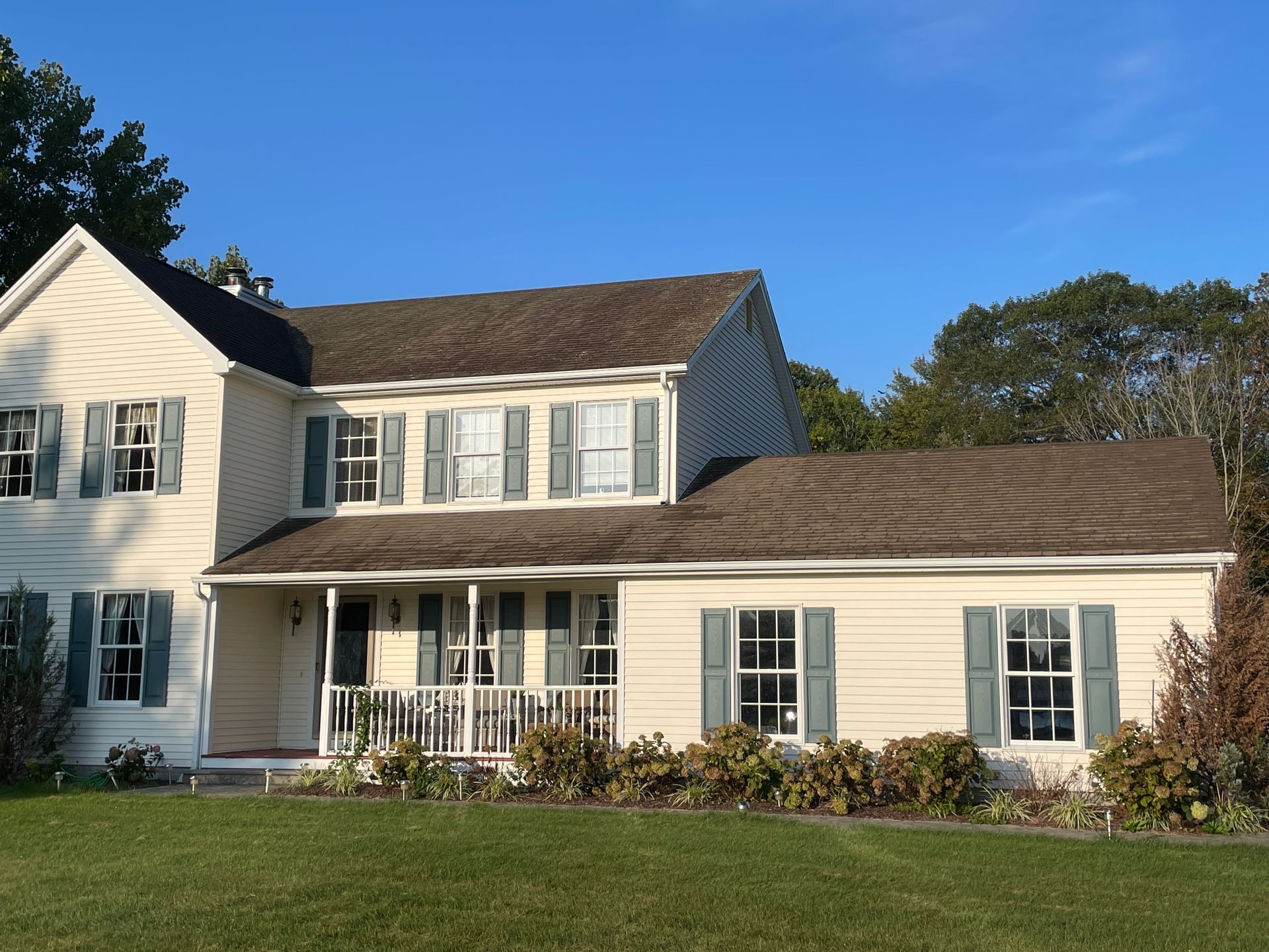 Beige two-story house with green shutters and a brown roof against a clear blue sky.