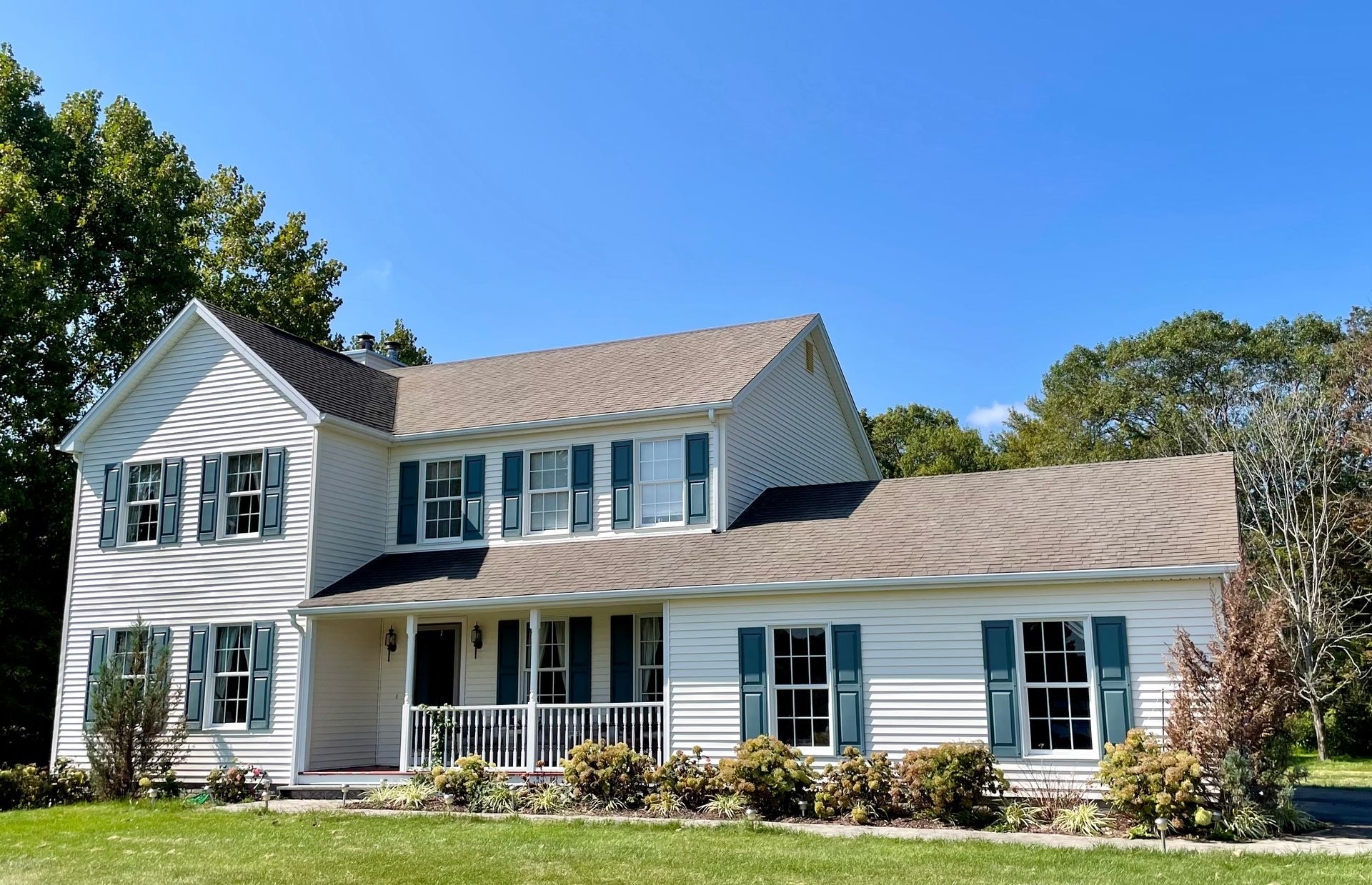 White two-story house with green shutters, brown roof, and porch on a sunny day.