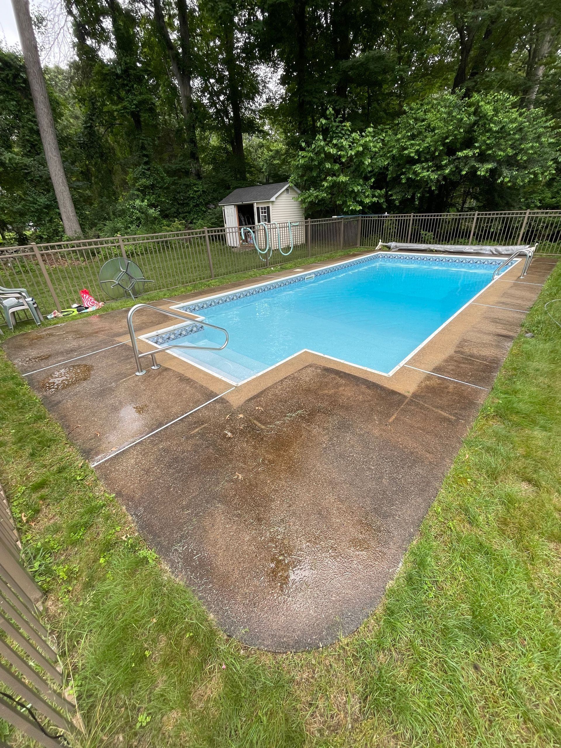 Rectangular swimming pool surrounded by concrete and grass, with a small shed and trees in the background.