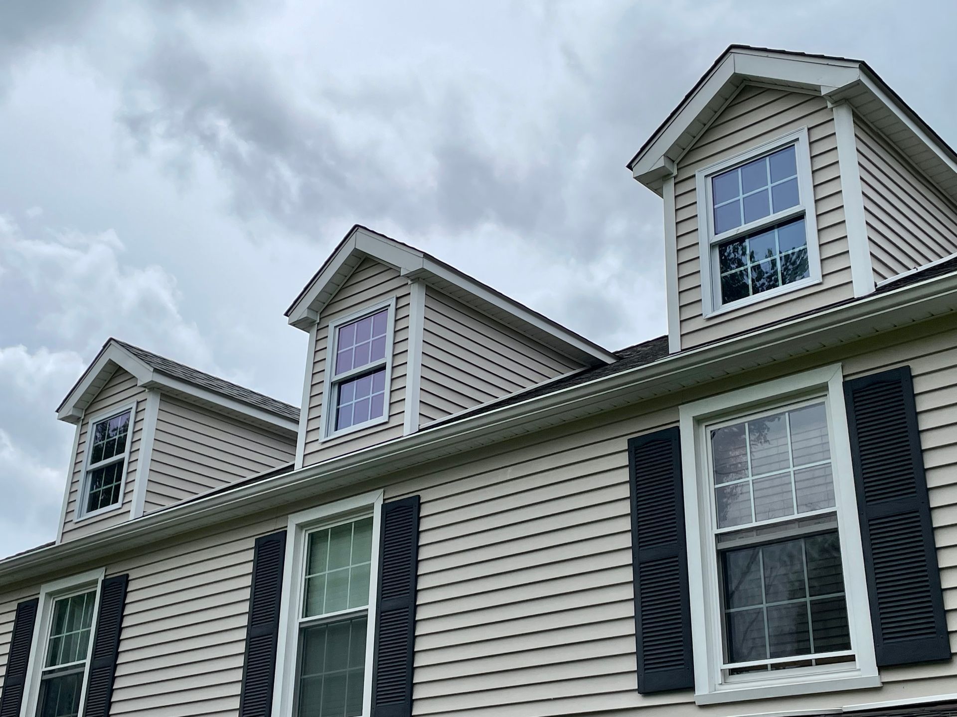 Beige house with dormer windows and black shutters against a cloudy sky.