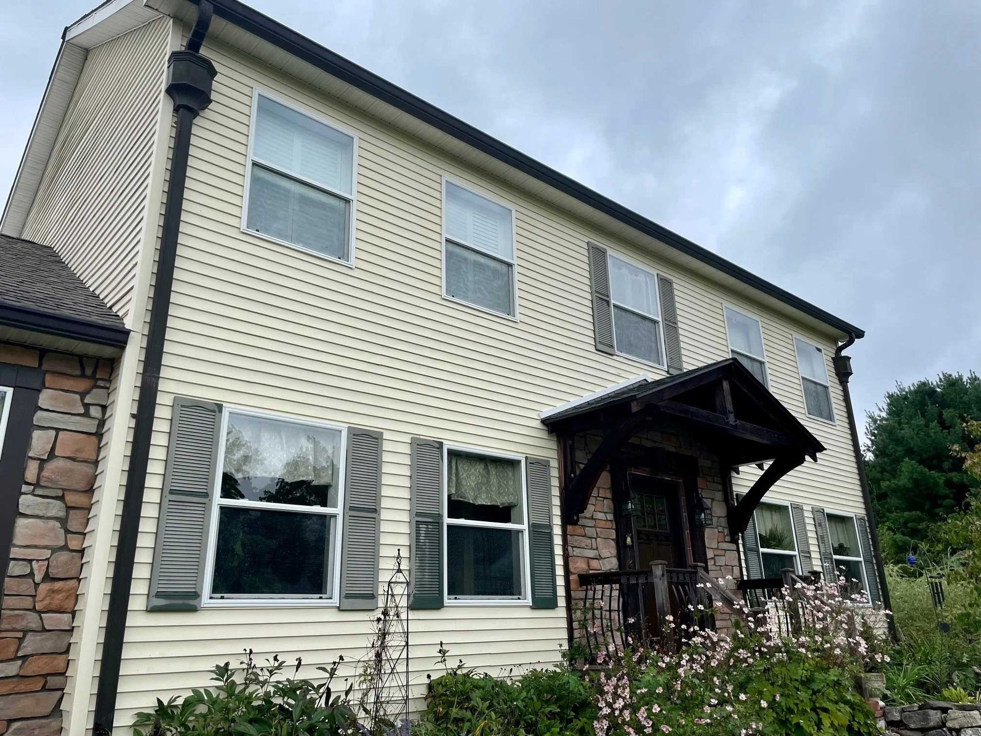 Two-story house with tan siding, stone accents, and dark shutters. Entryway has a porch and flowers. Overcast sky.