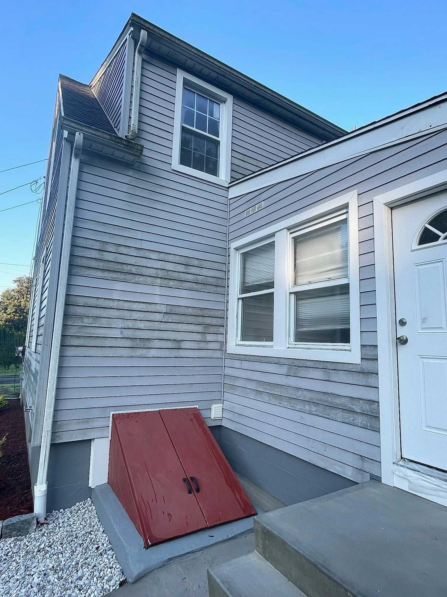 Side of a gray house with a red basement door and a white front door, under a blue sky.