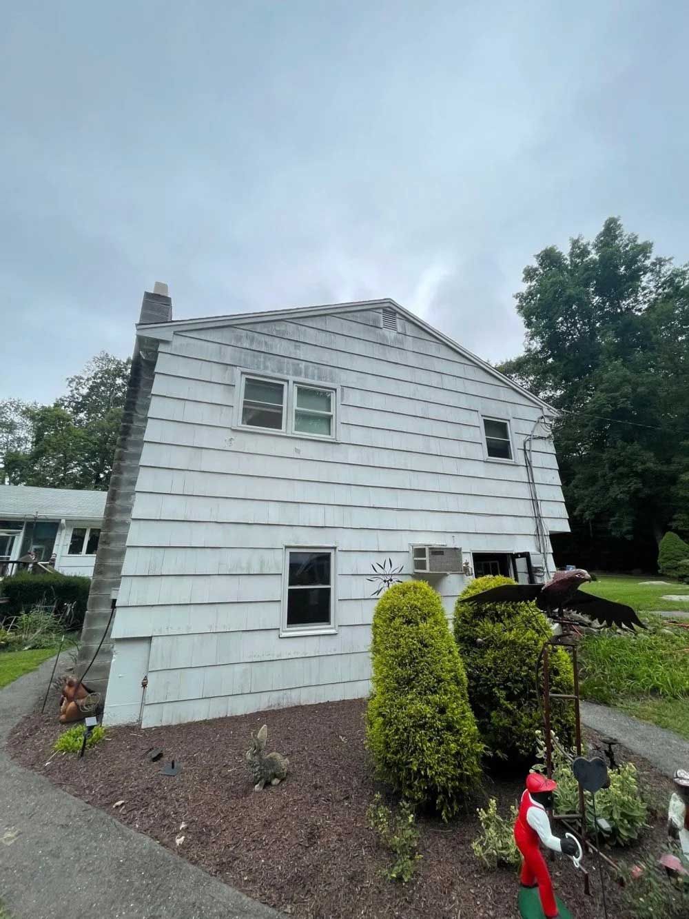 White-sided two-story house with two windows and an air conditioner. Garden with bushes and figurines in front.
