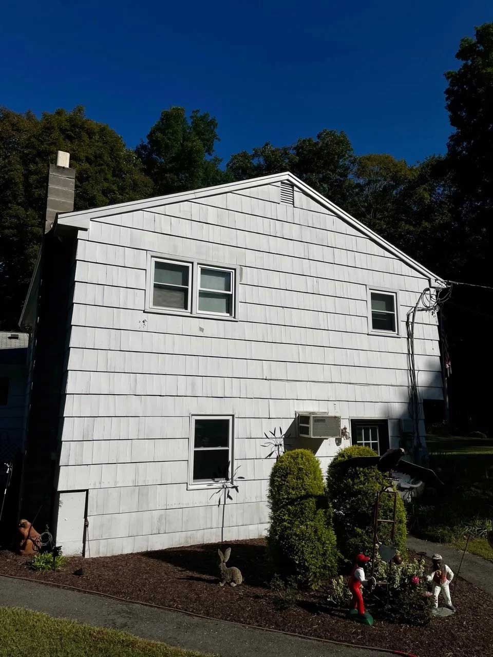 White-sided two-story house with gabled roof, windows, chimney. Landscaping in front, trees in the background, blue sky.