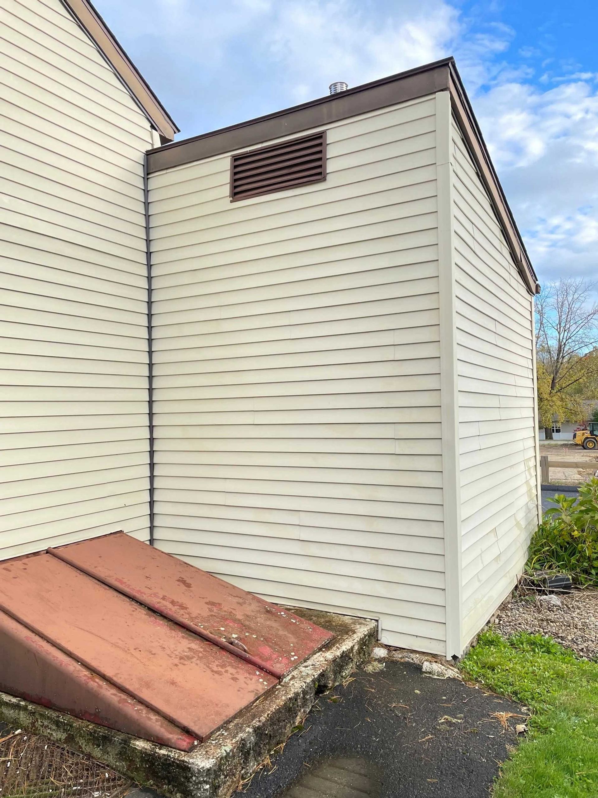 Side of a building with light siding. A rusty metal door is at the base. Brown vent near the roof.