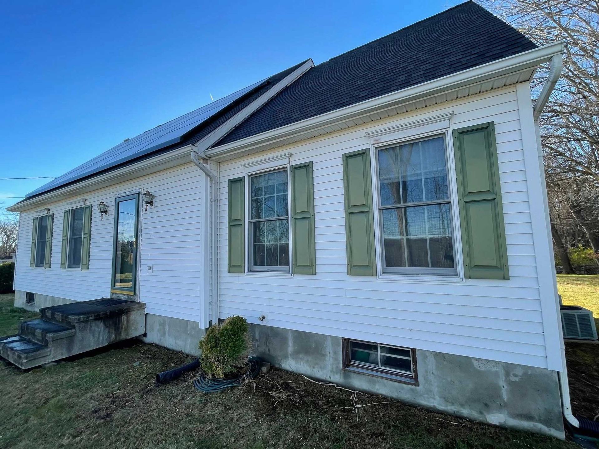White house with black roof, green shutters, solar panels, and a blue sky.