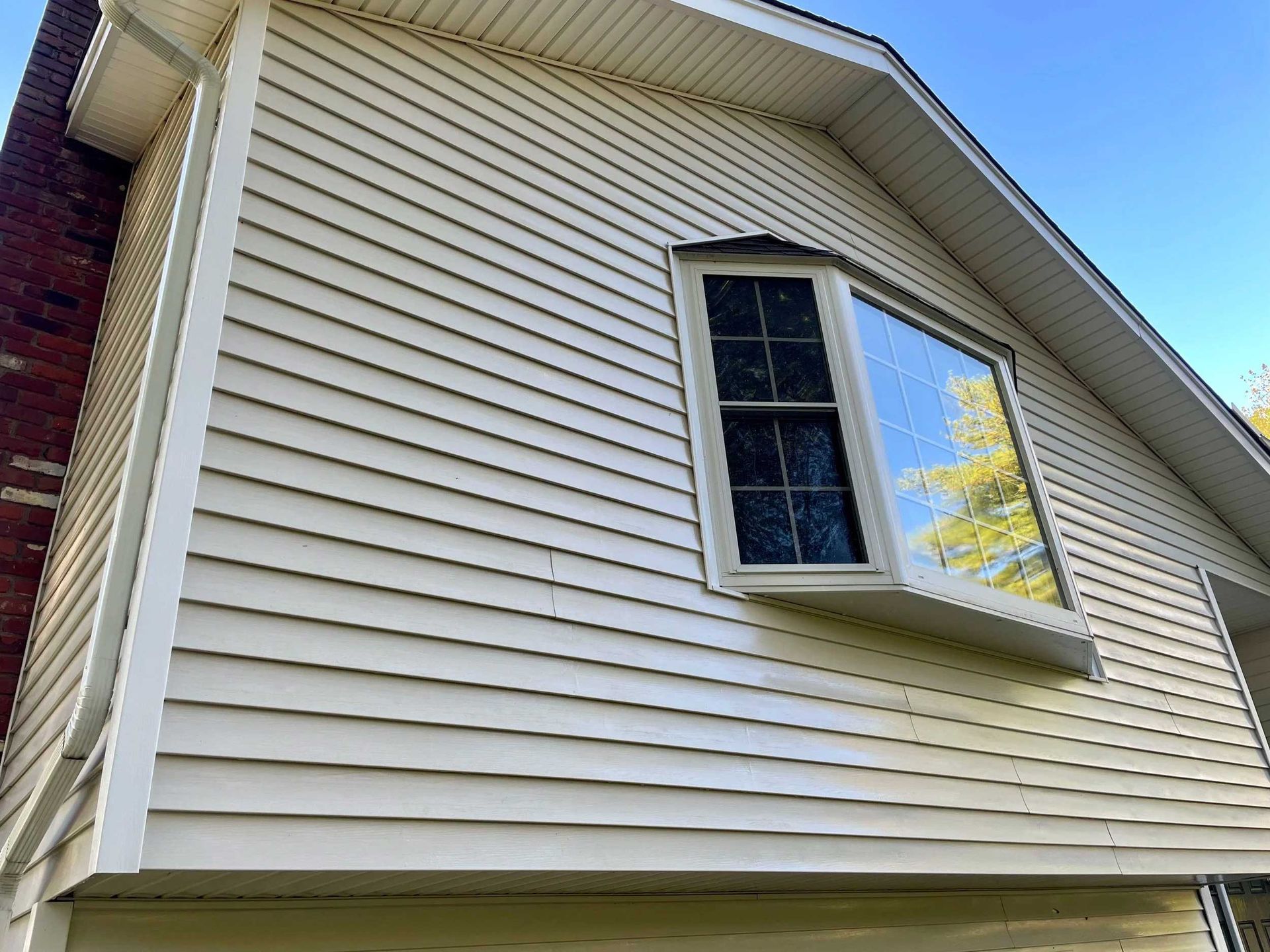 Tan siding on a house with a bay window reflecting the sky and trees. Brick chimney on the left.