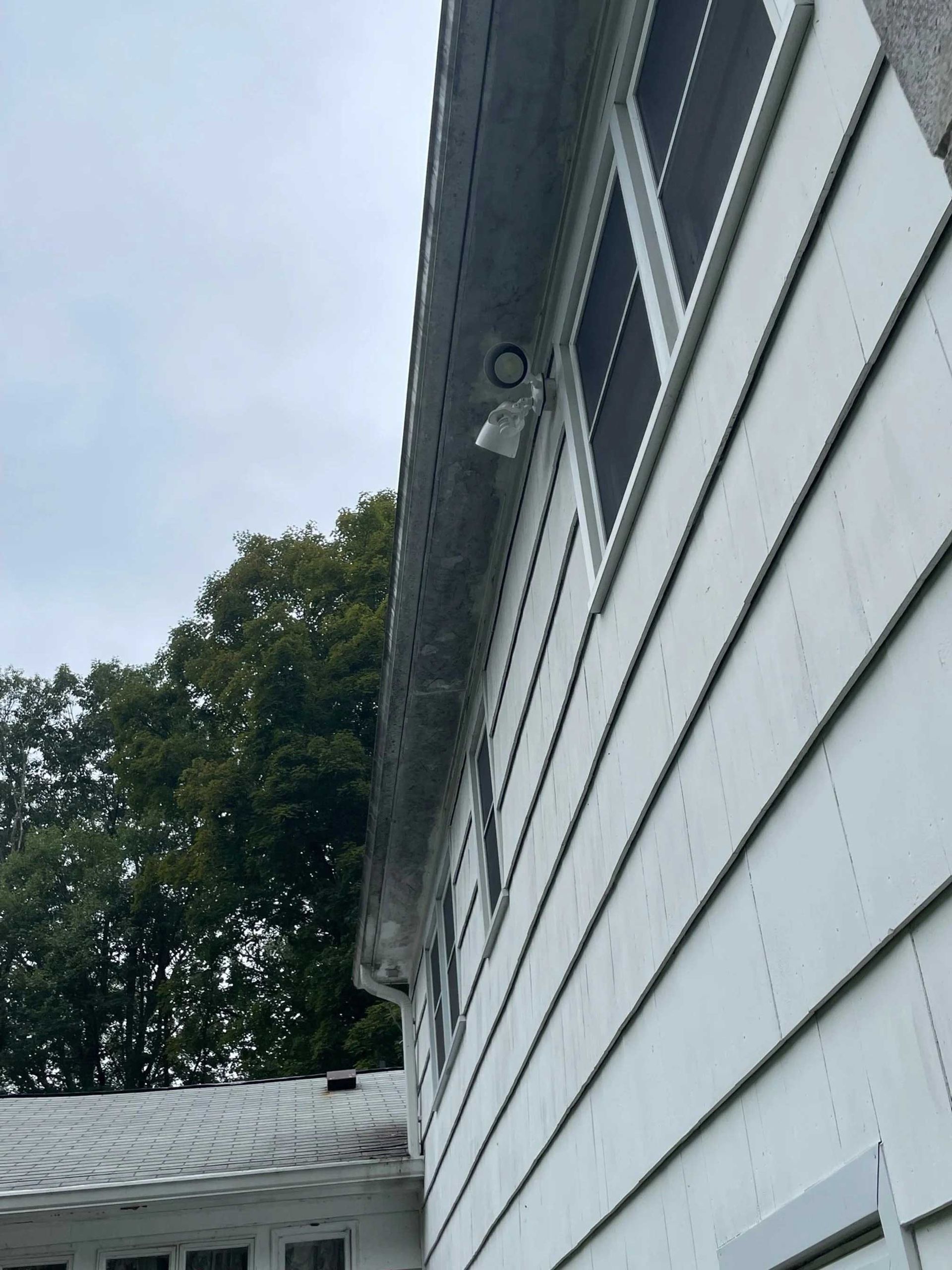 White house siding with a security camera mounted under the eaves next to a window. Overcast sky.