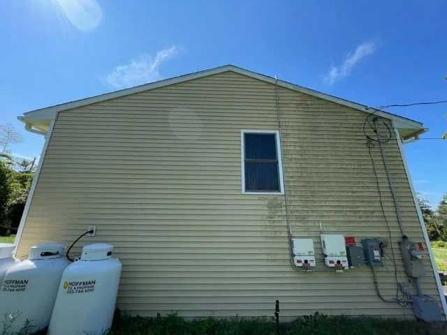 Tan-sided house with a single window and electrical boxes. Propane tanks on the left. Clear blue sky.