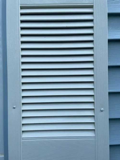 White louvered shutter against blue siding, mounted with two small fasteners.