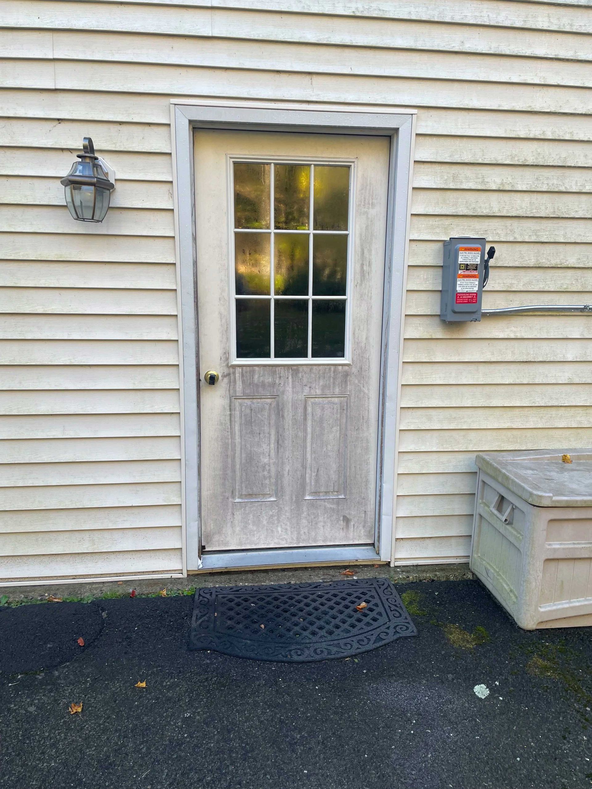 Exterior door with glass panes, worn white siding and trim. Dark doormat and electrical box.