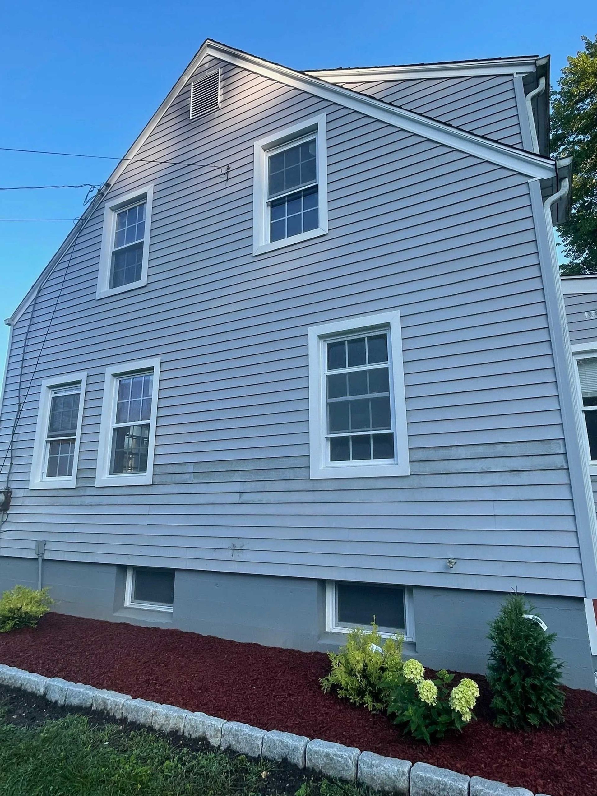 Side view of a two-story gray house with multiple windows and a red mulch-covered bed.